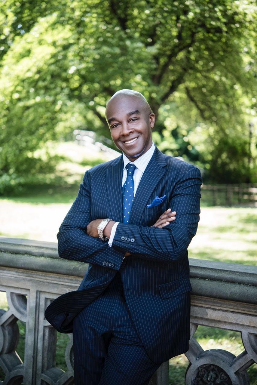 The color photograph shows Michael Carby, of Jamaican heritage leaning against a stone railing located outdoors. He’s wearing a blue pin stripped suit, with a blue polka dot tie, a blue handkerchief and a watch. His arms are crossed and he’s smiling at the camera. It’s a sunny day with green grass and green trees and bushes in the background.