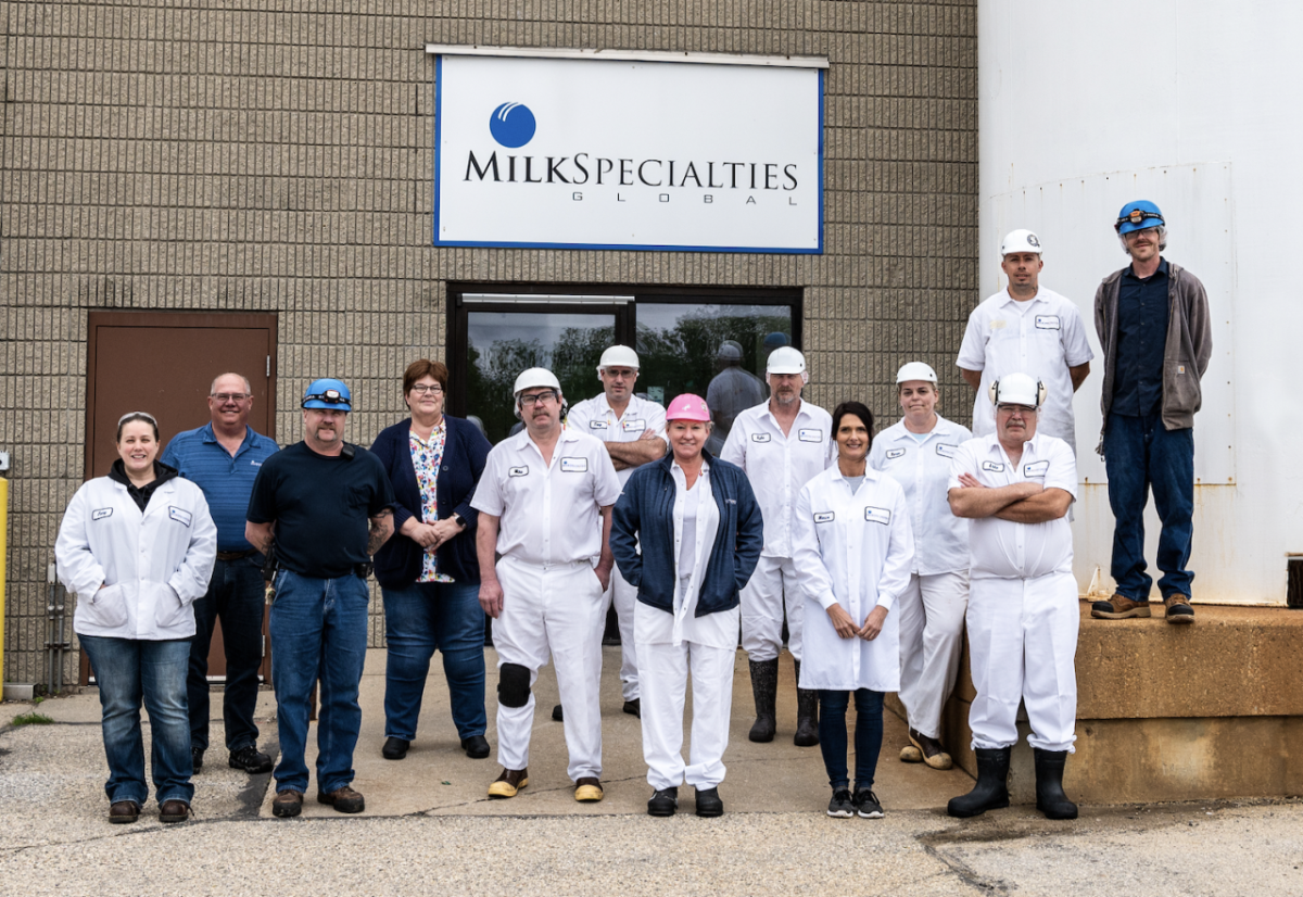 A small group of people posed outside a building. Some in all-white uniforms. "Milk Specialties" sign behind them. 