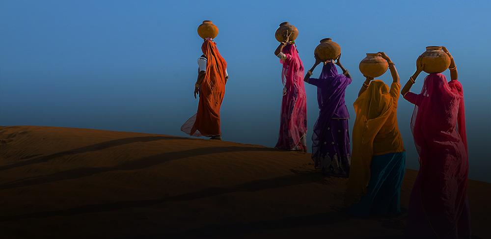 Women carrying pots of water across a desert.