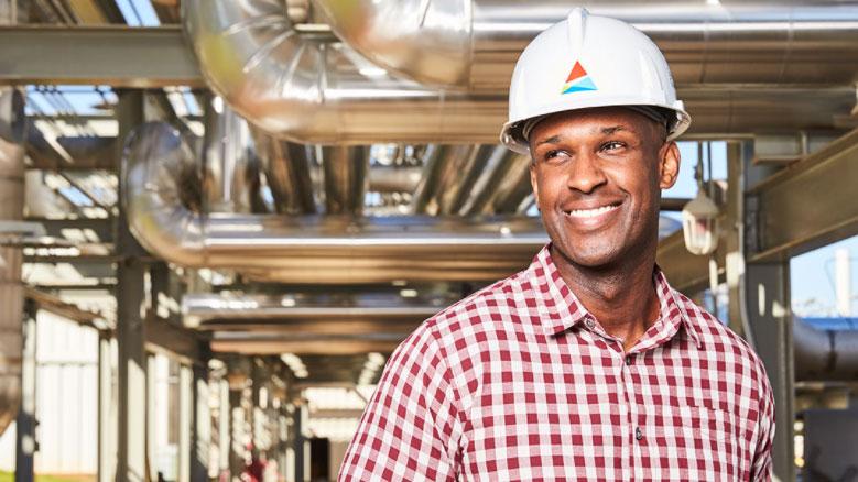 A person in a hardhat with southern company logo, underneath industrial piping. They are smiling and wearing a white and red checked button up shirt.