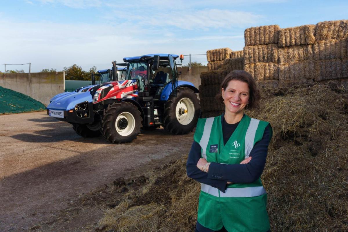 Person smiling with their arms folded, stood in front of a tractor and hay bales 