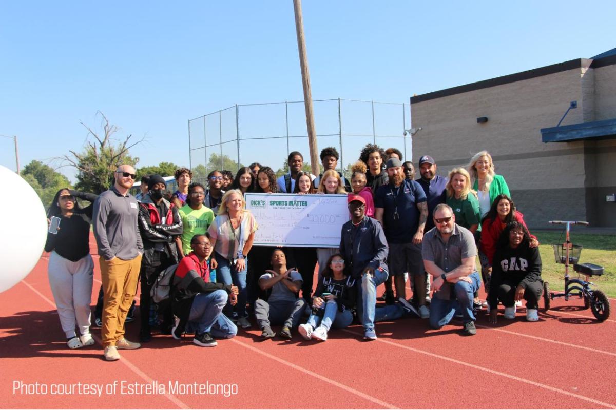 A group of people on a running track, some holding a large check. "Photo Courtesy of Estrella Montelongo" in the corner.