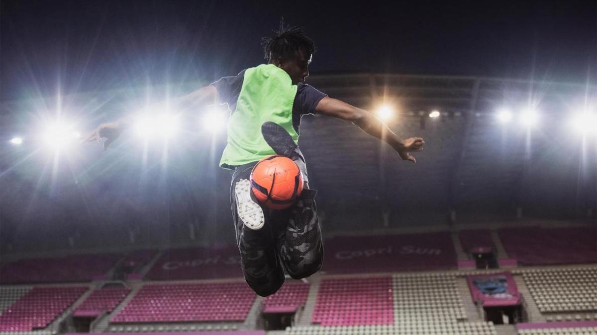 The back of a person doing a soccer trick move with the ball between their feet mid air. A lit stadium in front of them.