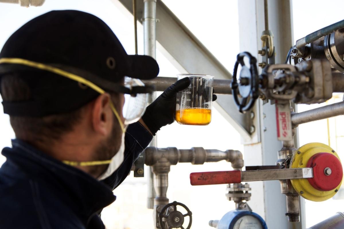 a person wearing protective glasses, mask and gloves holding a beaker of clear yellow liquid. Industrial pipes and valves in front of them.