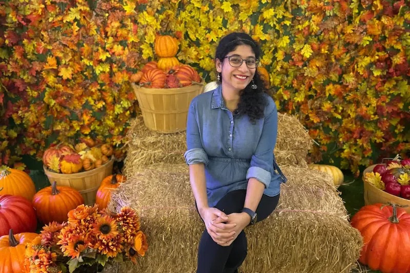 Sanjana seated on a bale of hay with pumpkins around her.