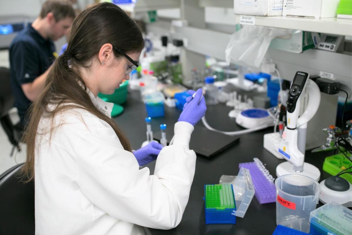 A person in protective lab gear working with pipettes and small containers at a table.