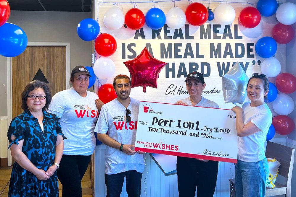 Group of people stood in front of a wall with red, white and blue balloons holding a Kentucky Fried Wishes funding cheque