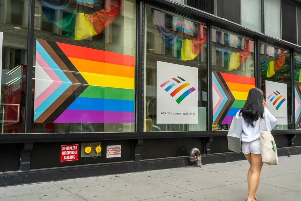 Pride flags shown in the windows of business on a city street. A woman is walking down the street with a shopping bag.