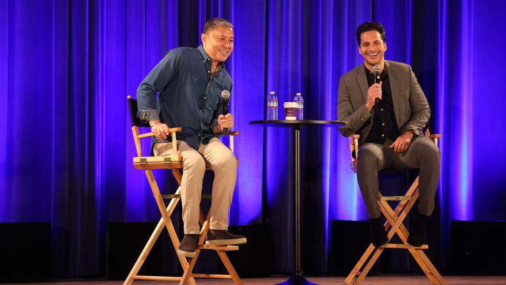 Two people on a stage in directors chairs, holding microphones.