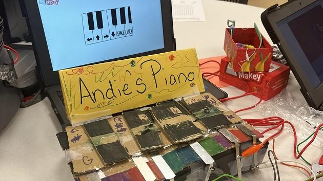 A computer screen with piano keys displayed. "Andie's Piano" on a sign below it and handmade controller in front.