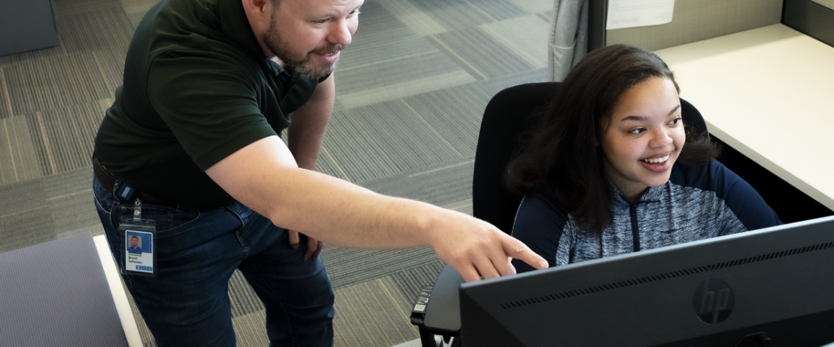 Person sat at a desk in front of a computer screen with someone stood next to them pointing at the monitor