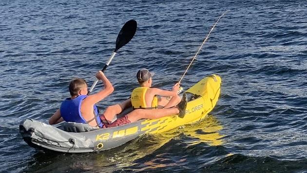 Two people in an inflatable kayak on a body of water, one with a fishing pole.
