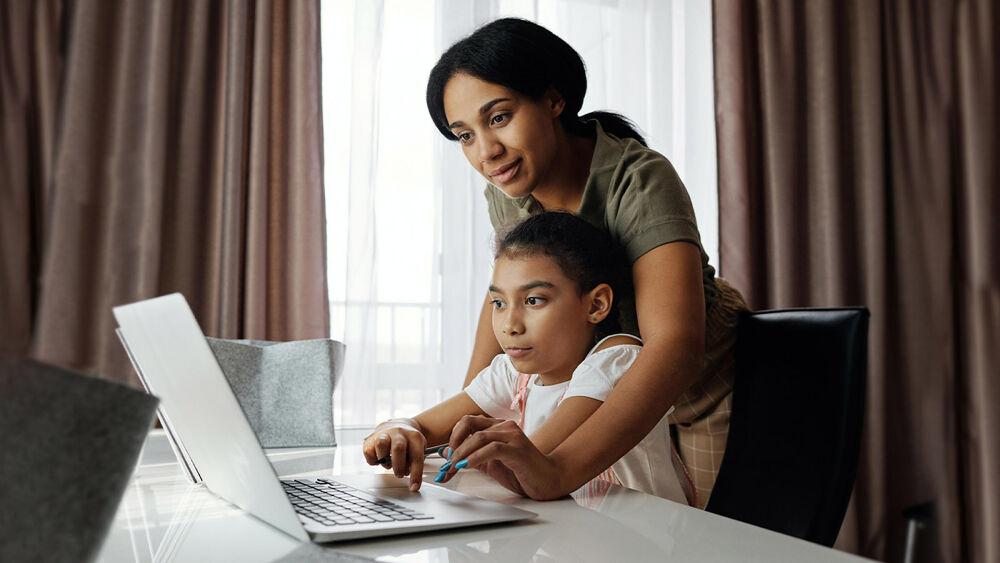 An adult leaning over a child, assisting them in using a laptop.