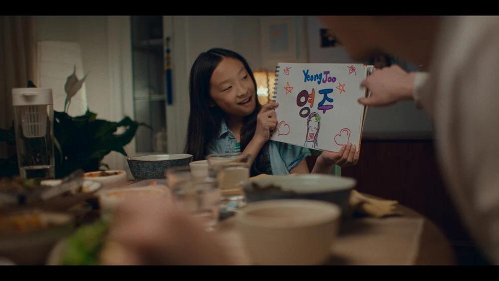 A child at a dinner table holding a notebook with hand-colored symbols, pictures and the name "Yeong Joo." An adult points to it.