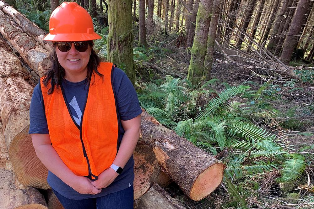 Tracie in front of cut timber in a forest wearing a safety hat and vest.