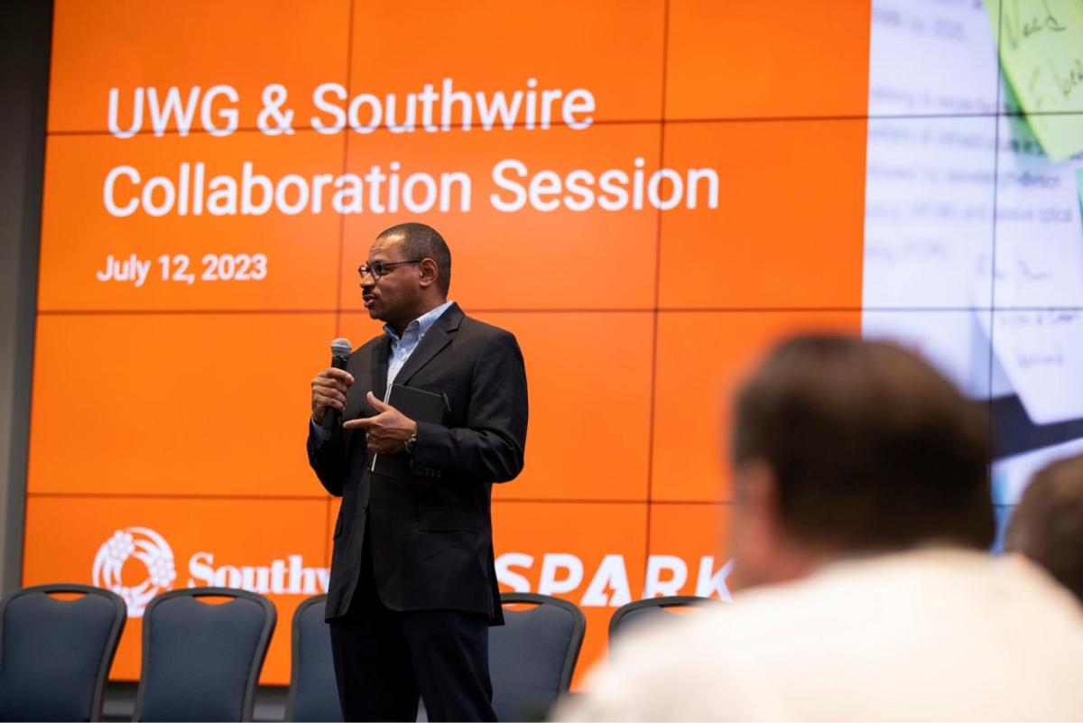 A speaker in front of a room of seated people. A digital screen behind with "UWG & Southwire Collaboration Session" 