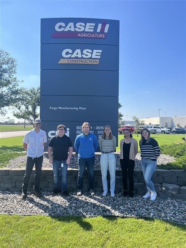 Six people posed outside in front of a tall sign for Case.