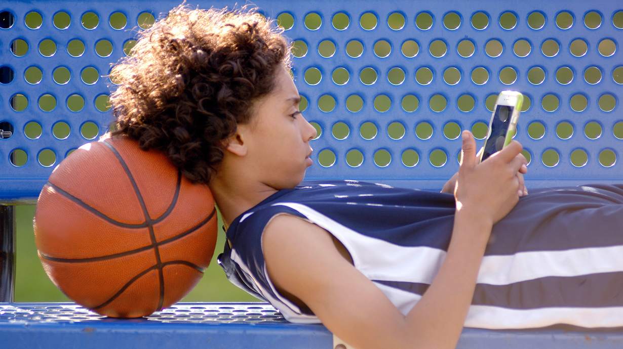 Child laying on a bench, phone in hand, head supported by a basketball