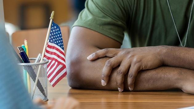 Close up of a person in army green shirt with arms crossed on a desk, a pencil holder with American flag and pens to the left.