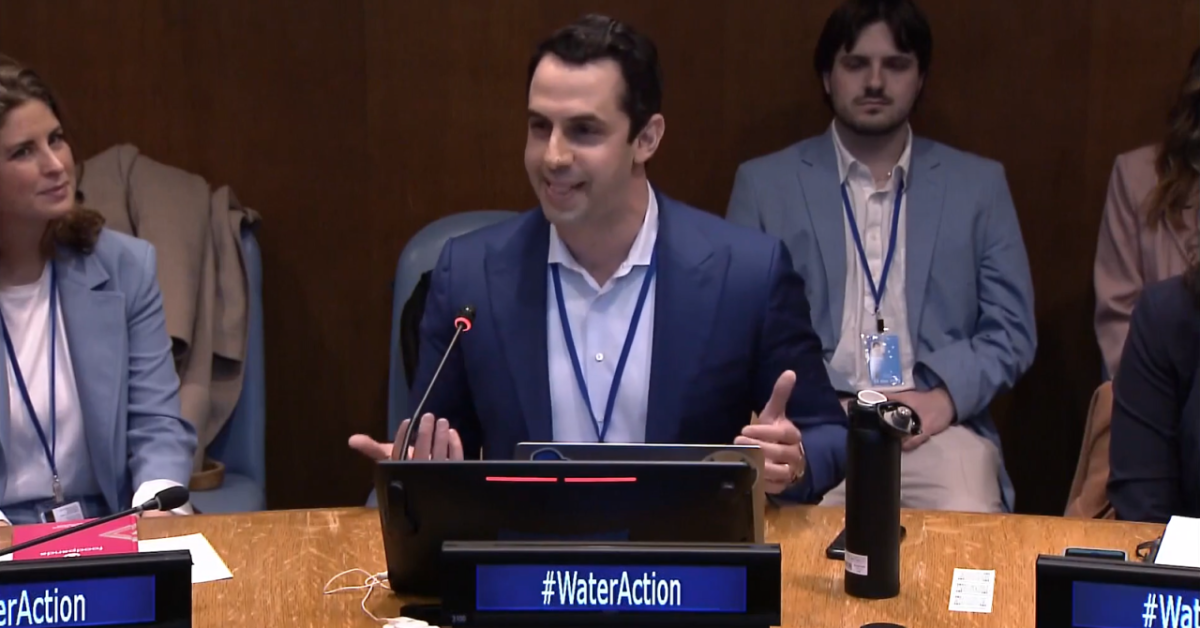 A person seated at a circular table with microphone and placard "#wateraction" Others to their sides and seated behind them.