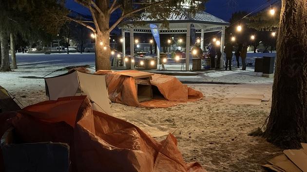 Makeshift shelters outside on a snowy ground. People gathered by a gazebo with string lights.