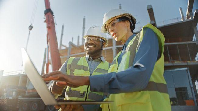 Two workers wearing a safety hat and hi-vis while looking at a laptop