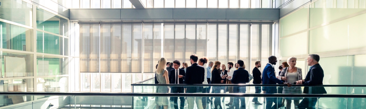 Group of about 25 business-attired people stand in small groups on a balcony in an office building with lots of outside light through glass windows