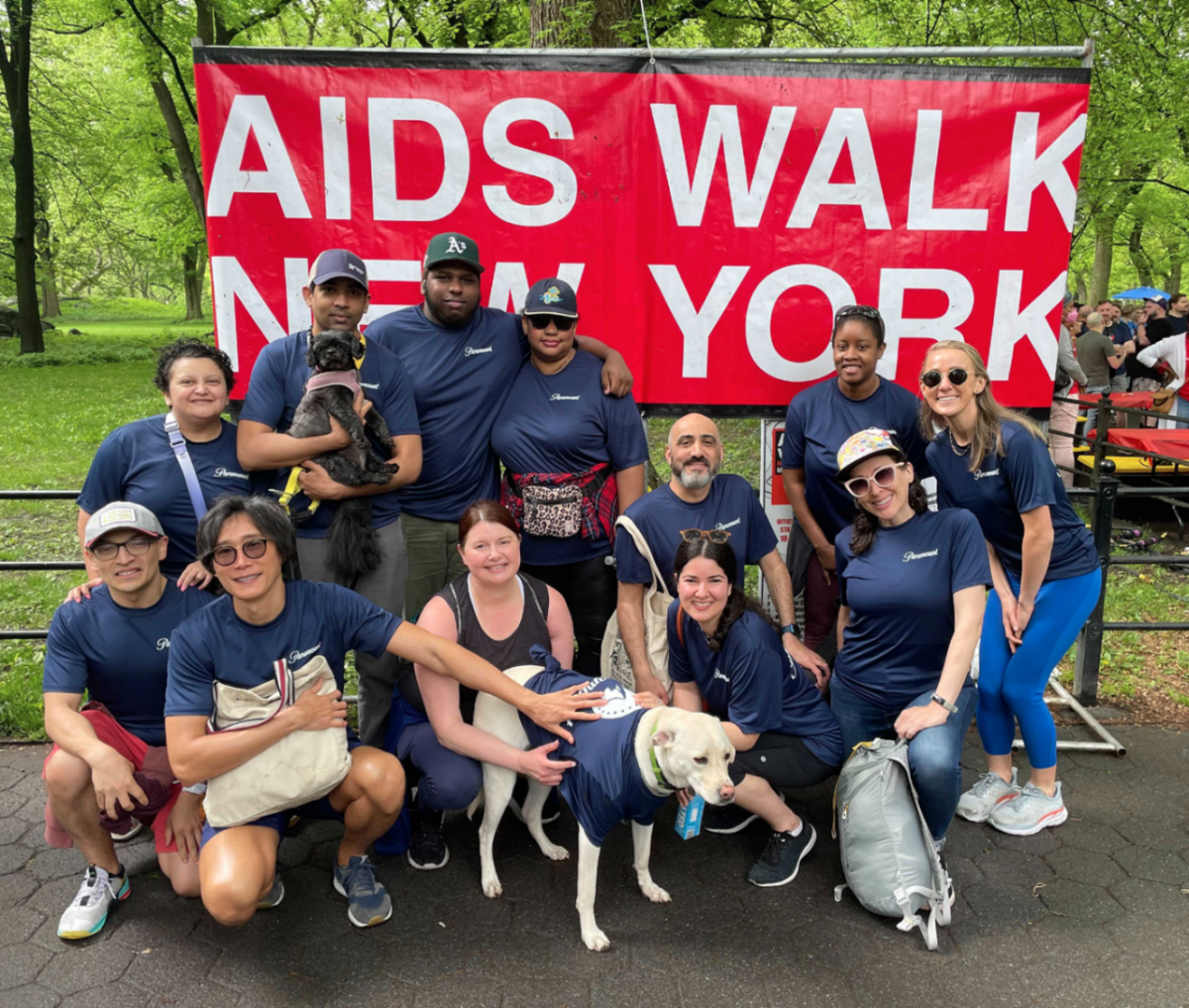A group of people, some sitting in front of others standing. Two dogs in front getting pet. All wearing matching t-shirts (even the good dog). "AIDS WALK NEW YORK" on a banner behind them.