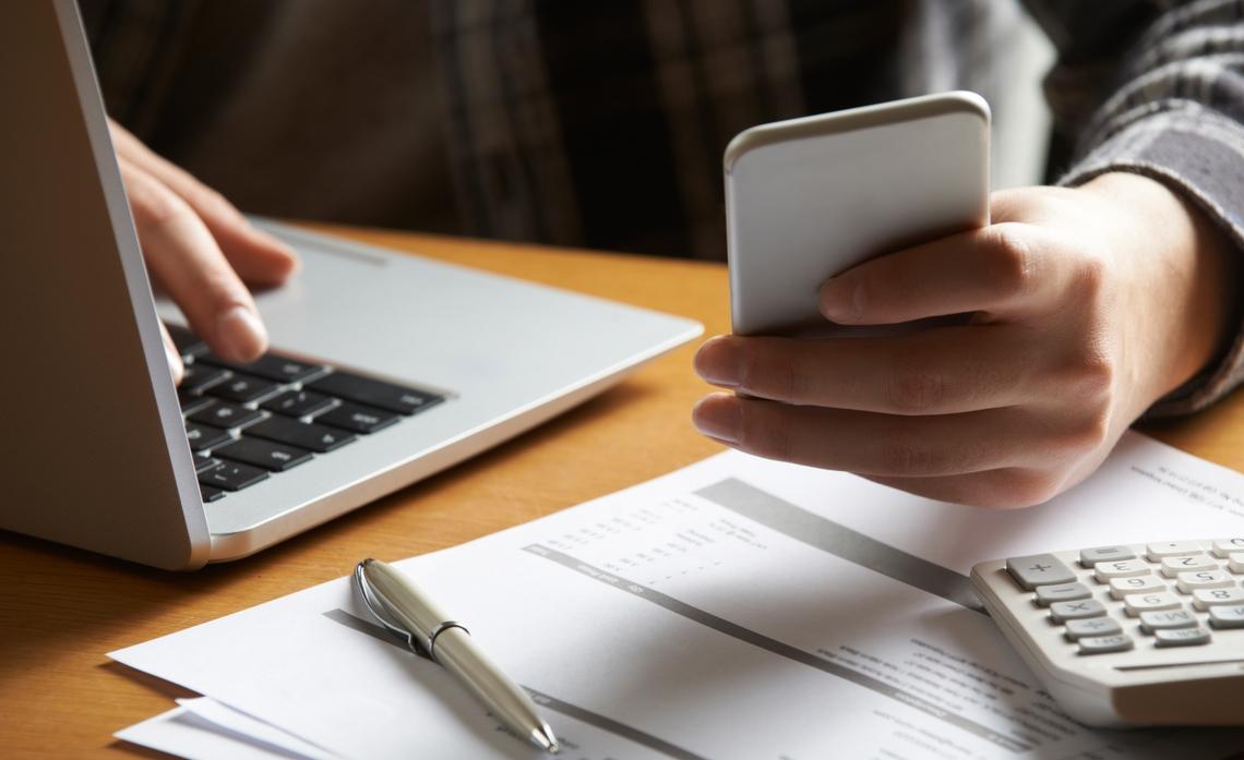 A person using a phone and laptop. Papers, a calculator and pen on the table too.