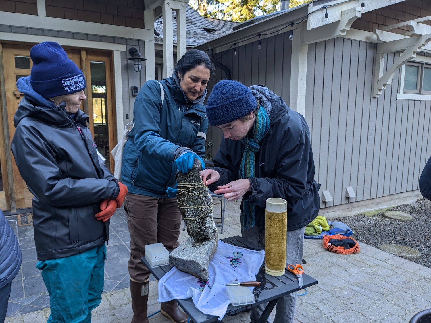 Three Puget Sound Restoration Fund team members prepare for kelp gardening by securing a thin rope around a rock.