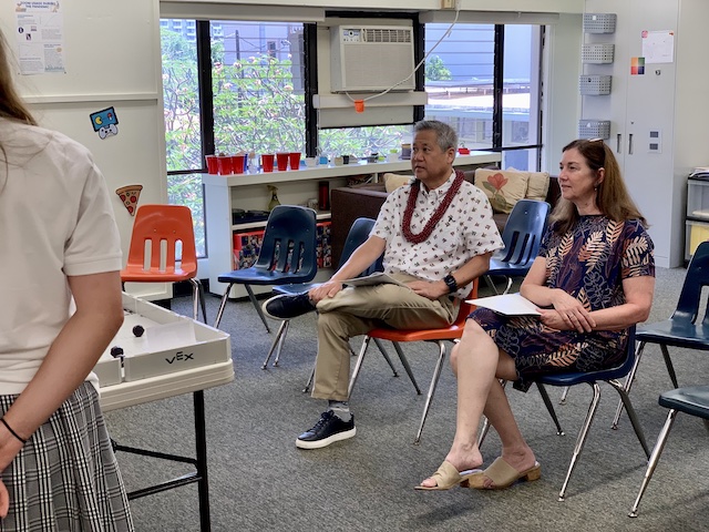 Two adults sitting in school chairs taking notes on presentations.