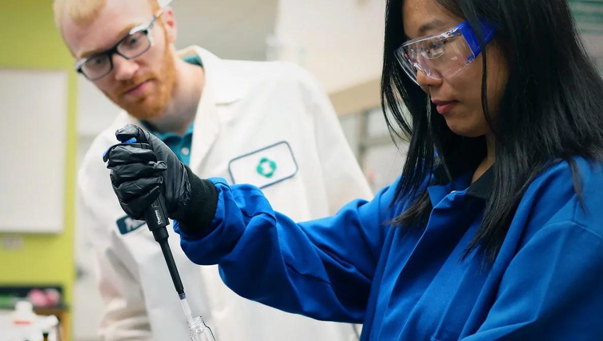 Two chemists, one observing another using a pipette.