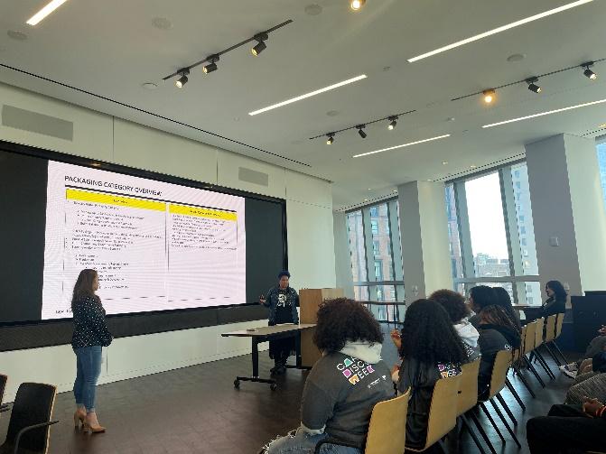 A classroom of students, two presenters at the front, a digital presentation behind them.