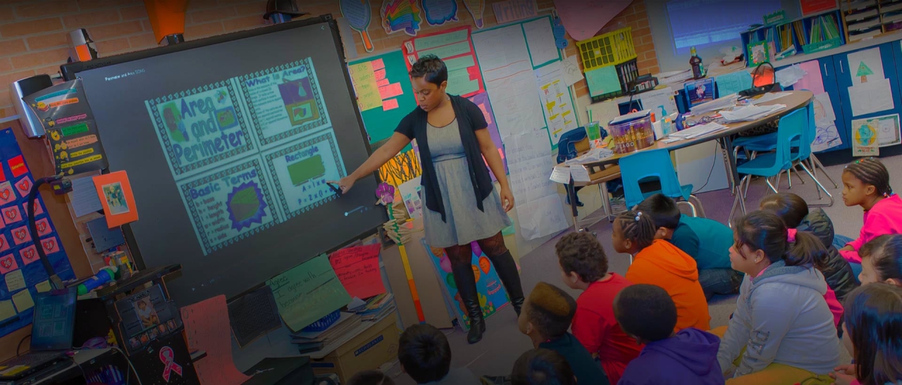 A teacher in front of a class of students, looking at a screen with lesson in area and perimeter on it.