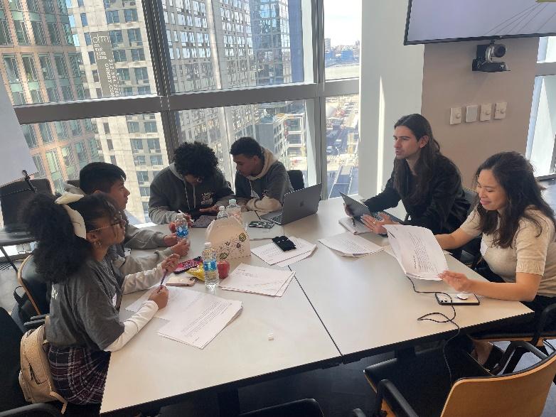 A group of people at a square table, papers strewn about. Large windows behind them.