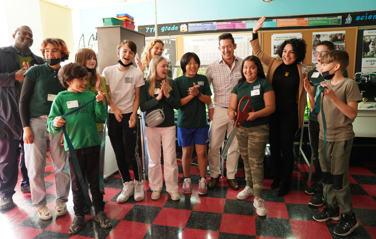 A group of adults and children are standing in a classroom clapping as a student cuts ribbon with oversized scissors. 