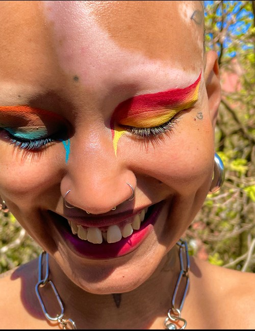 A person smiling with multiple piercings and colorful makeup