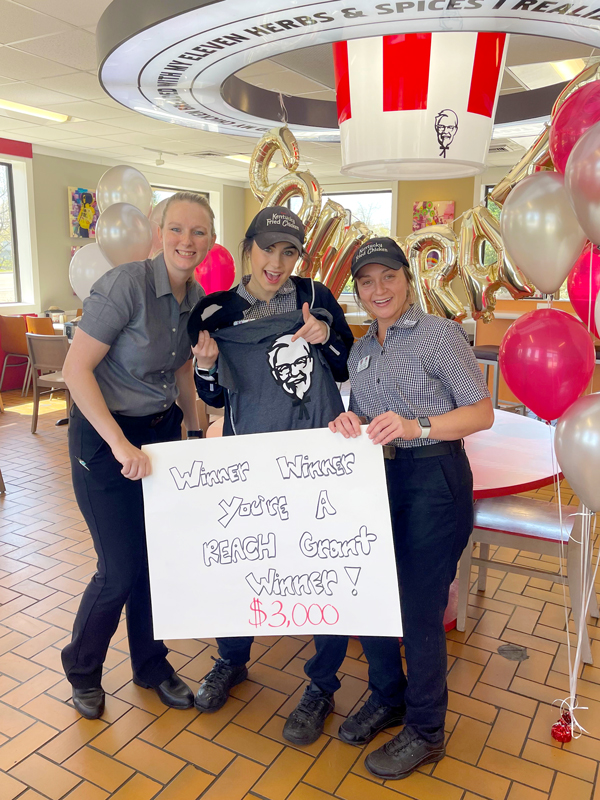 3 KFC employees hold a sign that reads " Winner, Winner. You're a REACH  grant winner! $3,000!"
