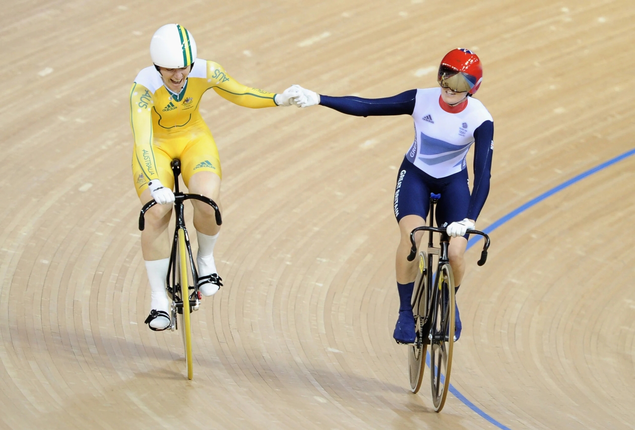 two cyclists on a wooden floor hold hands as they're riding