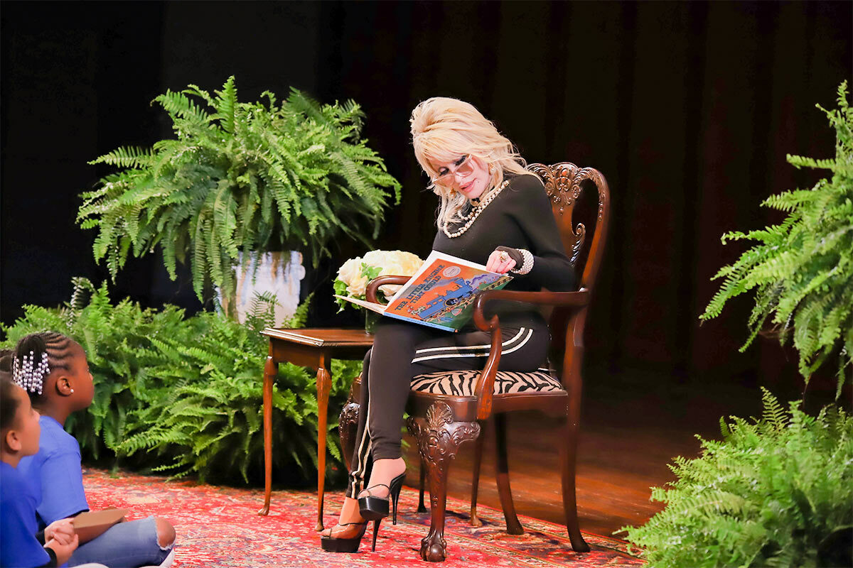 Dolly Parton sitting in a chair on stage reading a book, children in front of her