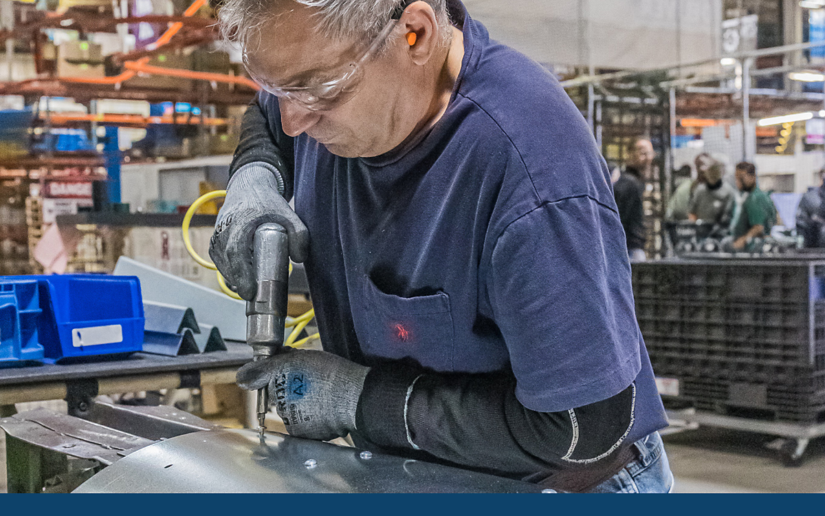 A worker in safety glasses and gloves drilling through a curved metal piece.