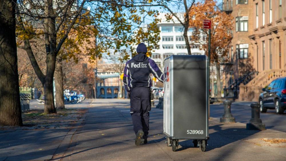 A person in FedEx uniform pulling a tall cart with FedEx Express logo on it down a sidewalk as a car goes by.