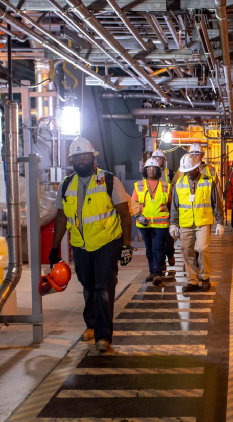 Group of five people on a marked walkway, each wearing high-vis vests, gloves and hard hats in an industrial setting