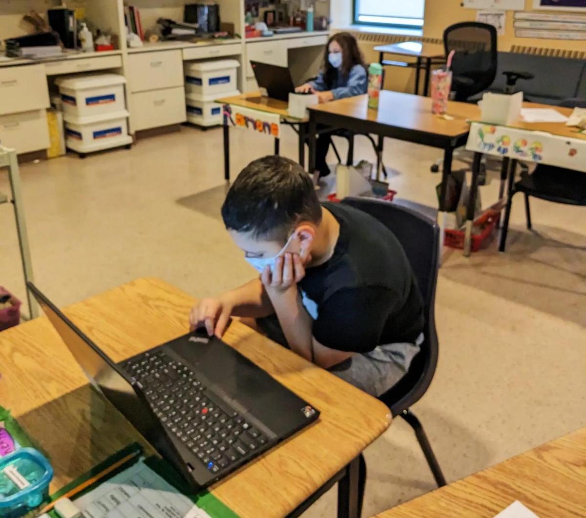 child in a mask and a black t-shirt sits at a desk with a lenovo laptop