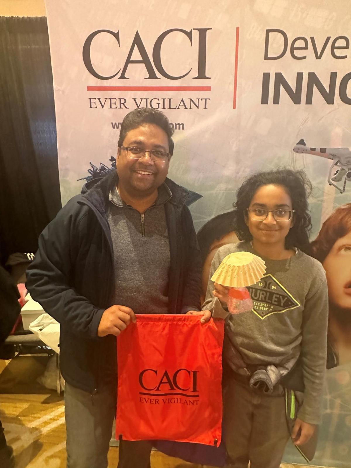 Two people in front of a CACI banner, holding up a CACI bag.