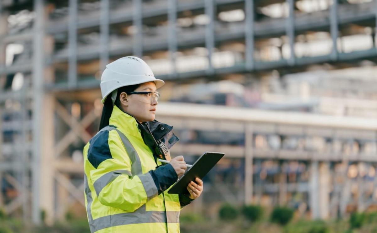 A person wearing high-vis clothing and hard hat, using a tablet. Large scaffolding behind them.