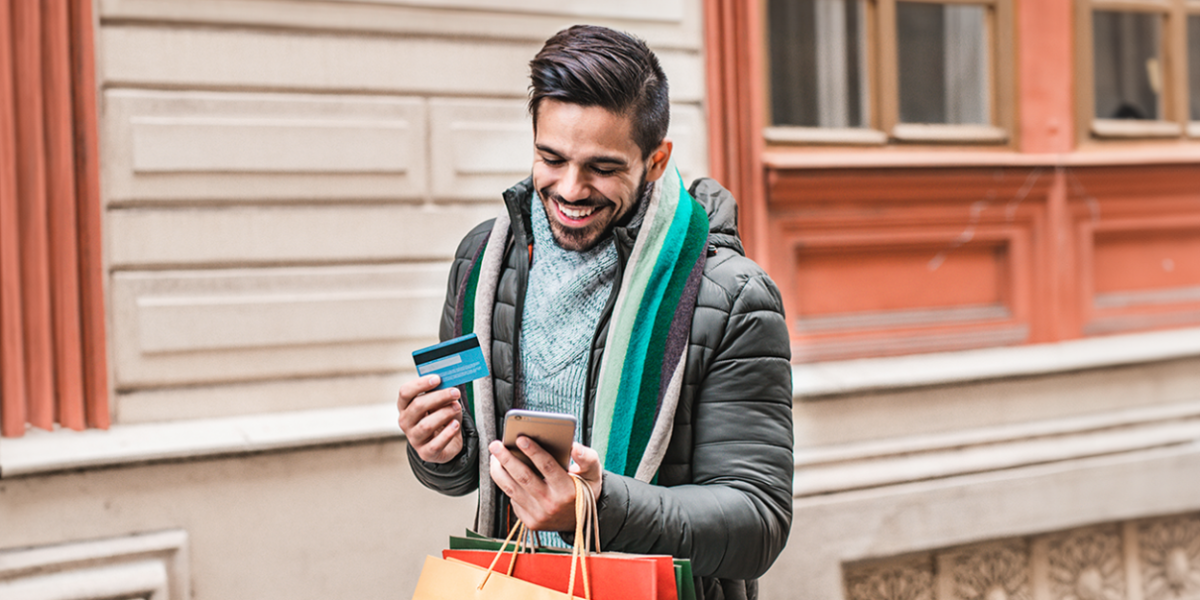 A smiling person outside holding a credit card and cellphone and shopping bags.