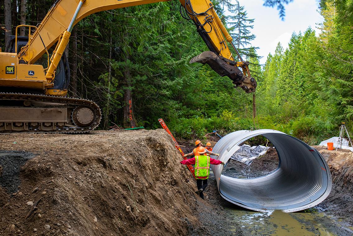 A large machine lifting a metal culvert as workers in a ditch position it.