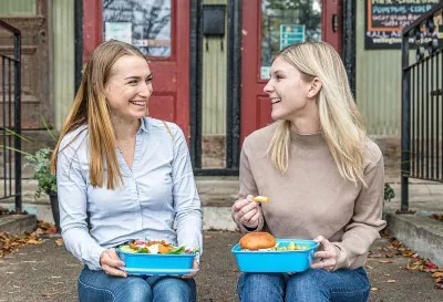 co-founders Jacquie Hutchings and Kayli Dale sitting on a front porch, eating out of blue containers