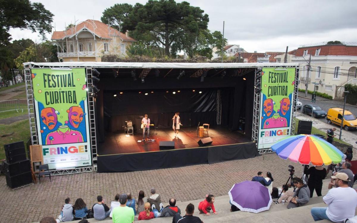 Two people performing on an outdoor stage, an audience in cement bleachers.